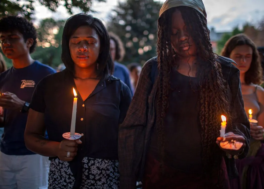 Multiple demonstrators at a vigil, with two young Black women at the foreground. Their heads are bowed and their eyes are closed. They each hold a lit, white candle, and a couple of people in the crowd behind them are also holding white candles.